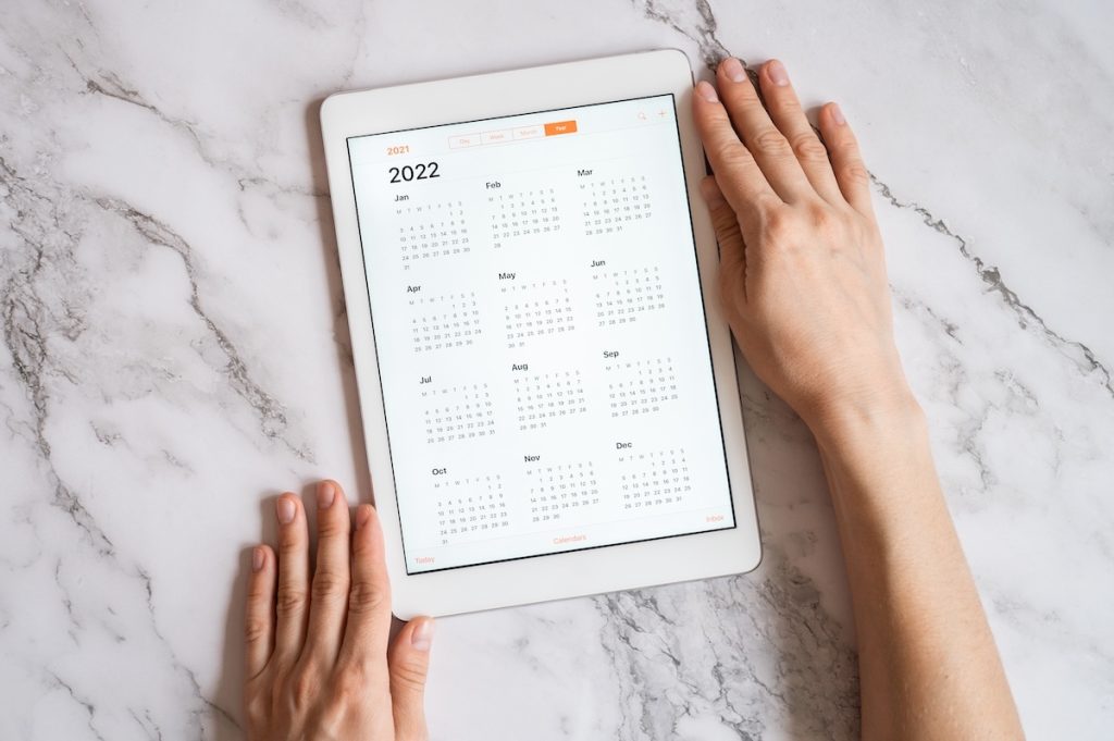 Tablet computer with an open app of calendar for 2022 year in a womans hands on a gray marble background. concept business or to do list goals with technology using. top view, flat lay